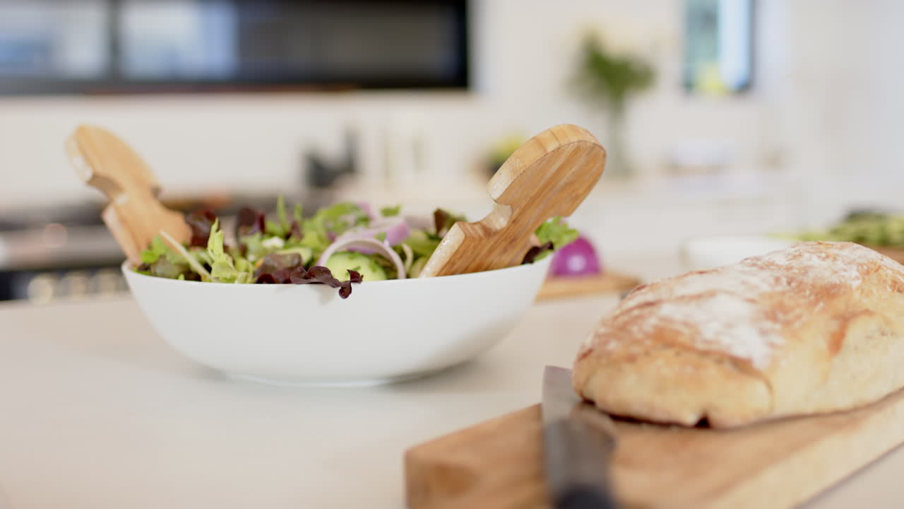 Preparing fresh salad in white bowl with bread on cutting board in kitchen