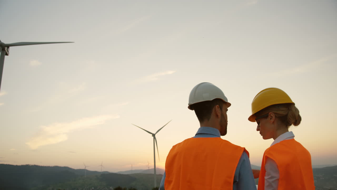Rear view of two Caucasian male and female engineers in helmets talking about the big windmills turbines