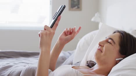 happy caucasian woman relaxing, lying in bed and using smartphone