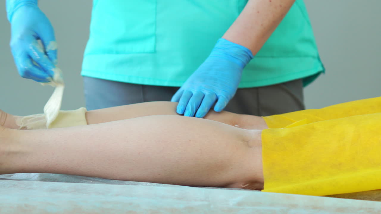 dos chicas en un salón de belleza maestro en una bata y guantes haciendo el procedimiento para eliminar el pelo en las piernas con una mezcla de azúcar