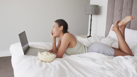 mujer biracial feliz acostada en la cama usando una computadora portátil y comiendo palomitas de maíz, cámara lenta