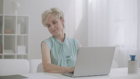 Una-Mujer-Jubilada-Con-Osteocondrosis-Estira-Las-Manos-Y-El-Cuello-Durante-El-Trabajo-Con-Una-Computadora-Portátil-Sentada-En-La-Mesa-Y-Escribiendo-En-El-Teclado