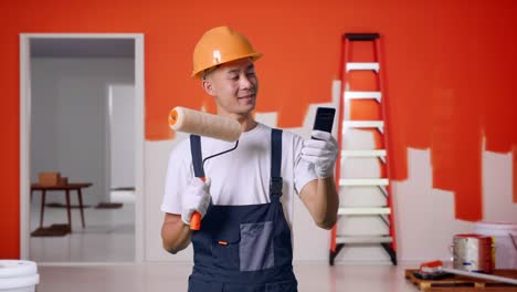 pintor asiático con casco de seguridad agitando la mano haciendo una llamada de video en el teléfono inteligente mientras está de pie con la pared de pintura en la habitación