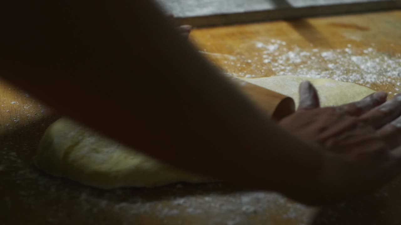 Freshly raised dough pressed flat using rolling pin on wooden kitchen top by chef, filmed as close up slow motion shot
