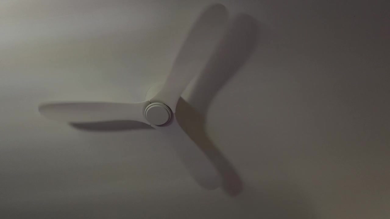 White wooden ceiling fan rotating slowly against a white ceiling in a dim room, minimal clean scene with soft shadows, calm motion and quiet atmosphere, simple interior detail, vertical composition
