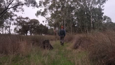 a fisherman walks through scrub and bush in the wimmera on a dry creek bed with a fishing rod in his backpack