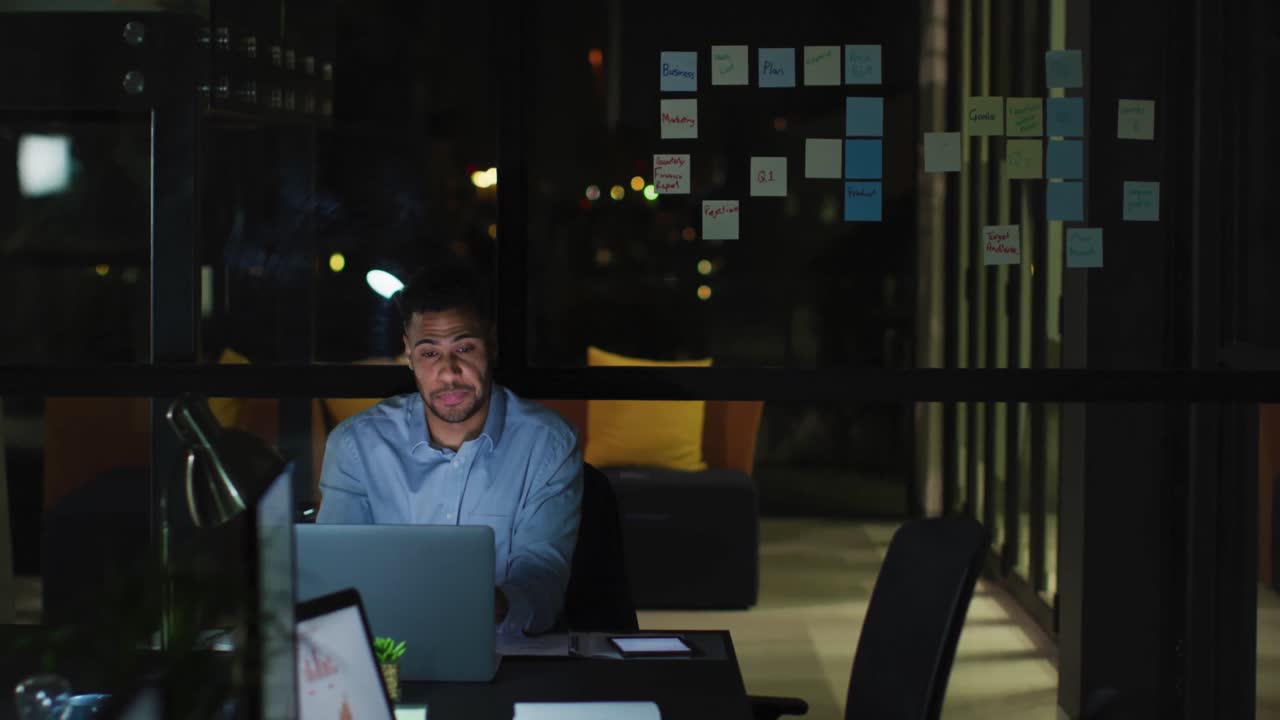 Video de empresario birracial cansado sentado en un escritorio usando un portátil, trabajando solo de noche en la oficina