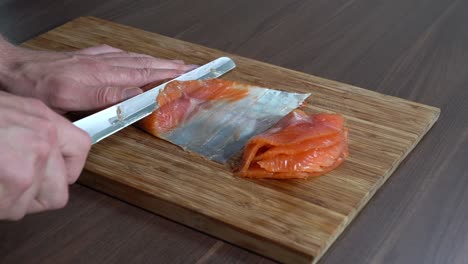 a male persons hand gently slicing smoked salmon on a wooden board with sharp knife - static slow motion