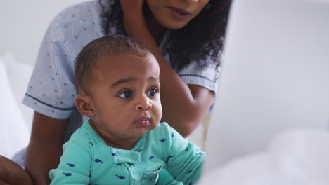 Mother-Wearing-Pyjamas-Cuddling-Baby-Son-In-Bedroom-Whilst-Working-From-Home-On-Laptop