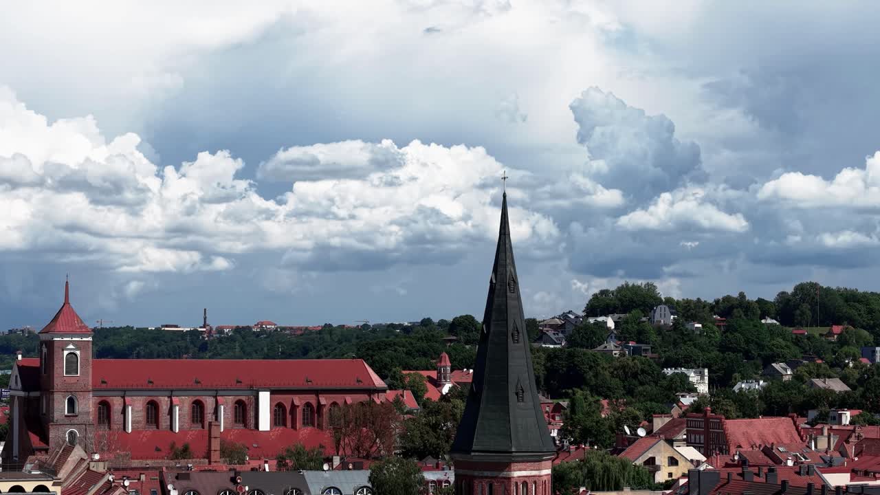 Scenic view of a European town with a tall church spire rising above red rooftops, surrounded by forested hills and dramatic summer clouds in the sky