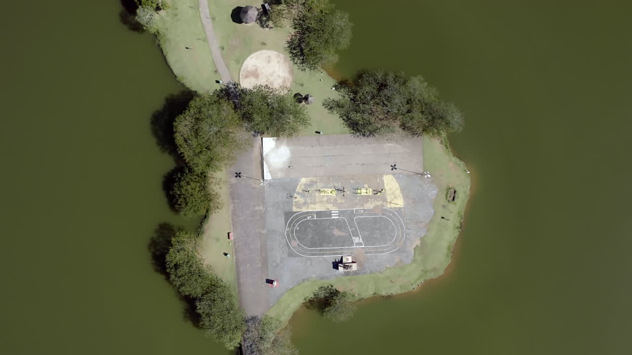 God's eye view shot of a little island in the center of the lake of a green park. Drone shot going up.