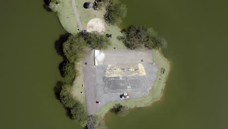 god's eye view shot of a little island in the center of the lake of a green park. drone shot going up.