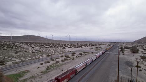 Imágenes-Aéreas-De-Drones-Del-Tren-De-Carga-En-El-Desierto-De-Palm-Springs-Con-Parques-Eólicos-En-El-Fondo,-Cacerola-De-Movimiento-Lento