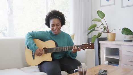 Fröhliche-Afroamerikanische-Frau,-Die-Zu-Hause-Auf-Der-Couch-Sitzt-Und-Gitarre-Spielt-Und-Singt,-Zeitlupe