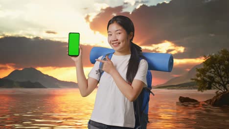 woman with backpack showing green screen phone at sunset lake