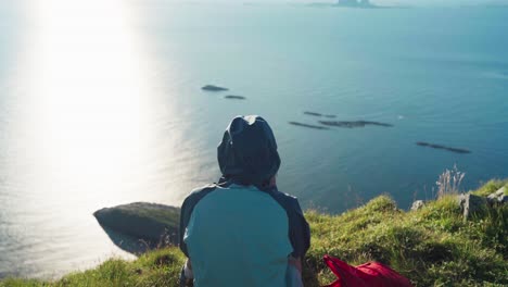 rear of a person in a hoody sitting on a the mountain edge in lovund island, norway