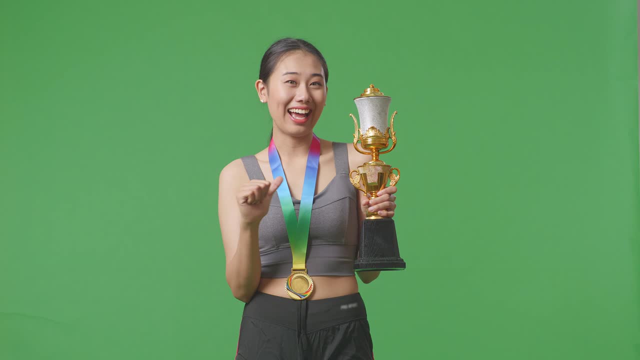 mujer asiática con una medalla de oro mostrando el trofeo de oro a la cámara, sonriendo, y señalándose a sí misma estando orgullosa de ganar como la primera ganadora en el fondo de la pantalla verde en el estudio