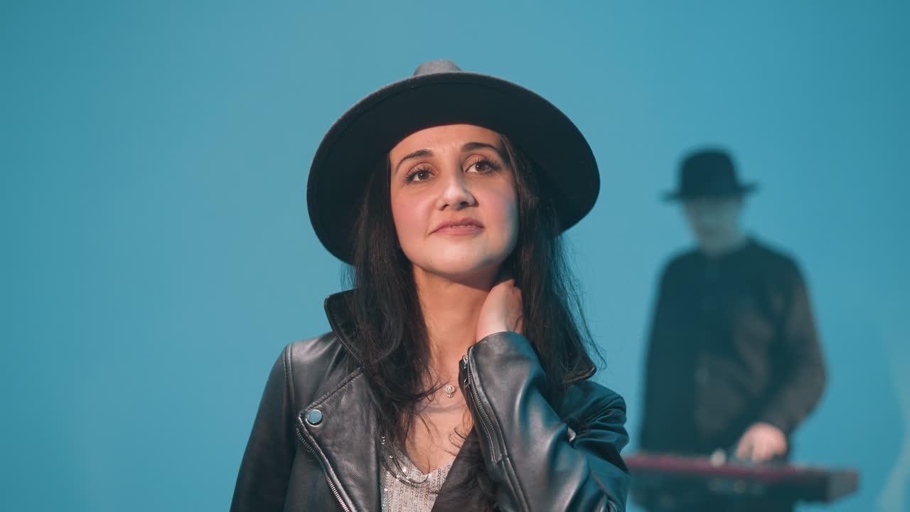 dama con una chaqueta de cuero negra y sombrero negro sonriendo mientras ajusta su cabello. en el fondo, un tecladista está borroso contra un telón de fondo azul, añadiendo profundidad a la escena