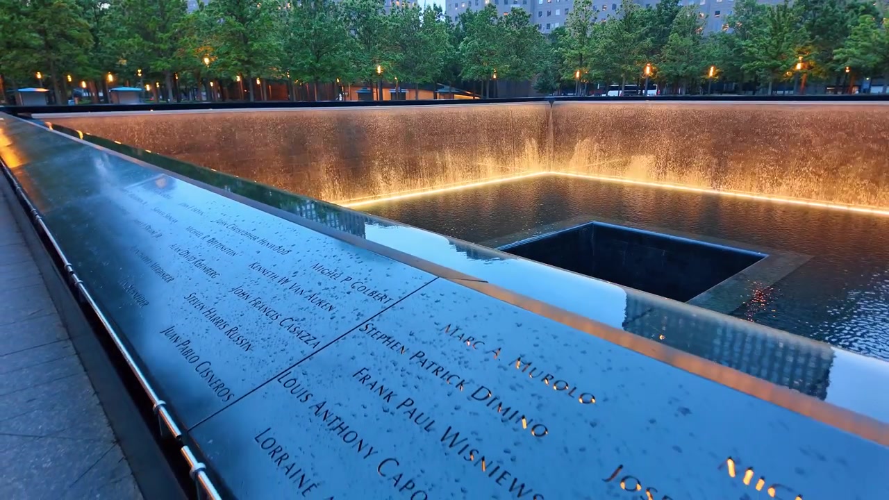 Indgraverede navne ved 11. september-mindesmærket, New York City, New York. Regndråber, tårnrefleksioner og et vandfald omgiver det mørke, firkantede tomrum.