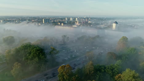 Toma-Aérea-Hacia-Atrás-Del-Castillo-Místico-De-Jelgava-Y-El-Tráfico-En-Las-Carreteras-Durante-La-Niebla-Voladora-Entre-La-Ciudad-De-Letonia