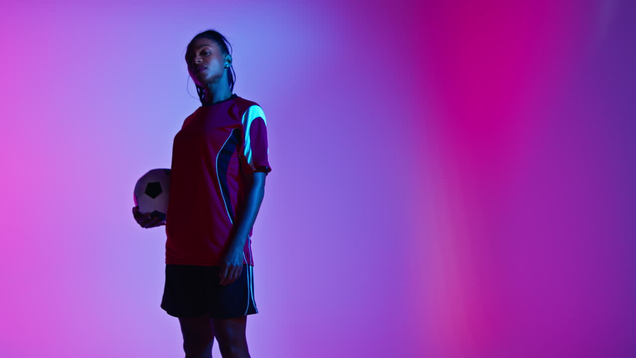 retrato de estudio de una jugadora de fútbol o fútbol femenina con la camiseta del equipo sosteniendo la pelota bajo el brazo disparado en clave baja contra una iluminación mixta colorida 4