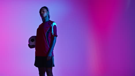 Studio-Portrait-Of-Female-Football-Or-Soccer-Player-Wearing-Team-Shirt-Holding-Ball-Under-Her-Arm-Shot-Low-Key-Against-Colourful-Mixed-Lighting-4