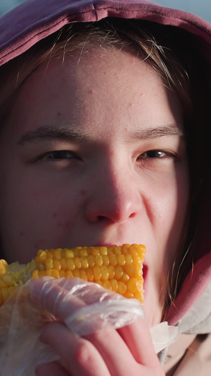mujer joven comiendo maíz en un palo envuelto en plástico blanco, vapor que se eleva desde el maíz fresco, luces bokeh en un fondo borroso, disfrutando de un bocadillo al aire libre en la noche de invierno con luces cálidas