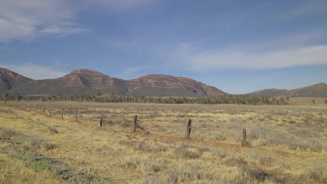 Panorâmica lenta sobre campos com vista para as Flinders Ranges na Austrália Meridional, com céu azul e montanhas vermelhas