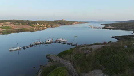 Drone-Desciende-A-Lo-Largo-De-La-Pared-De-Roca-Hasta-El-Muelle-De-Madera-De-Los-Barcos-Pesqueros-En-Sa-Nitja-Menorca-España