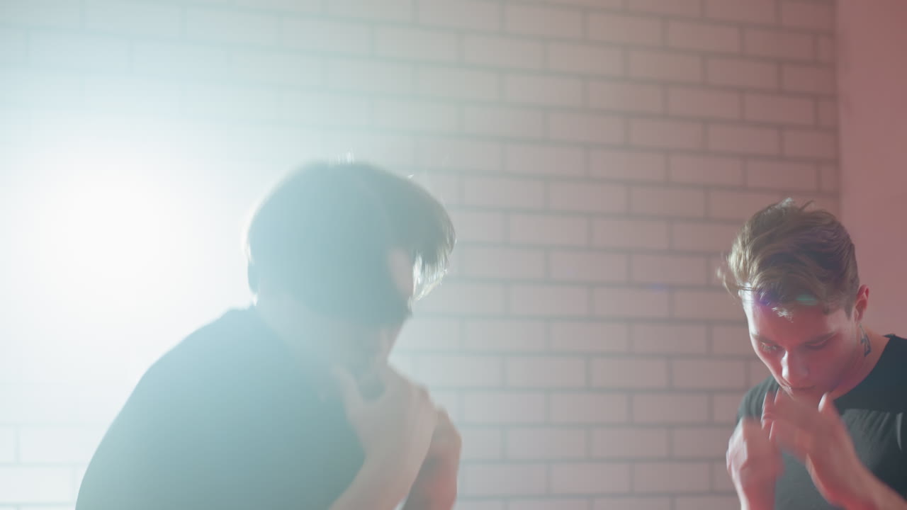 Boxer with short hair and visible tattoo on neck throwing hands in gym under strong backlight, showing exhaustion, pain, or reflection during training, expressing struggle,human emotion in boxing practice