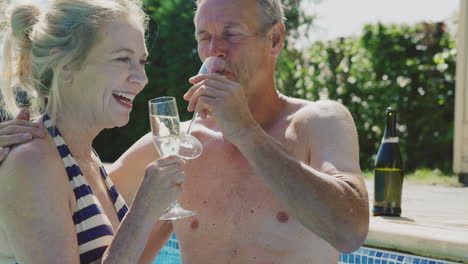 retired senior couple relaxing in swimming pool on summer vacation celebrating drinking champagne