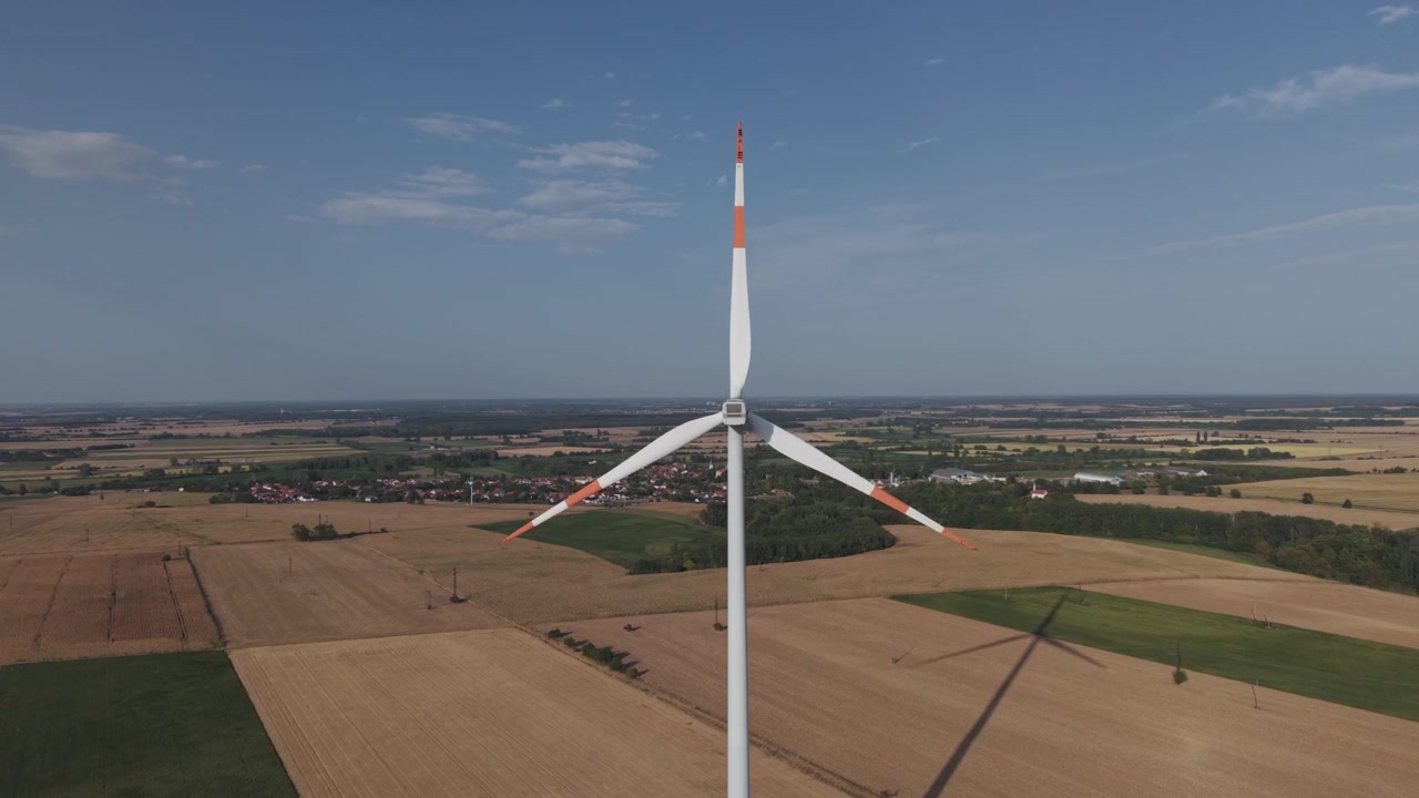 Aerial view of a large wind turbine rising above expansive agricultural fields, with a rural village and patchwork farmland in the background.