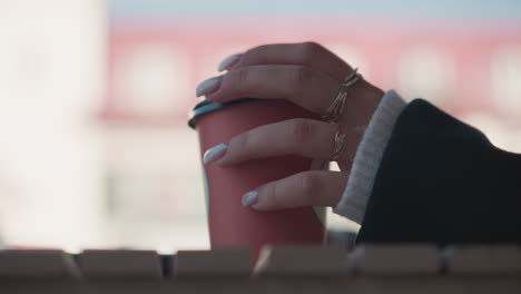 close-up of woman's hand with manicured nails holding red coffee cup while gently rubbing the lid, showcasing delicate jewelry and stylish rings against a blurred background