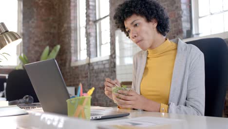 focused biracial casual businesswoman using laptop and having lunch in office in slow motion