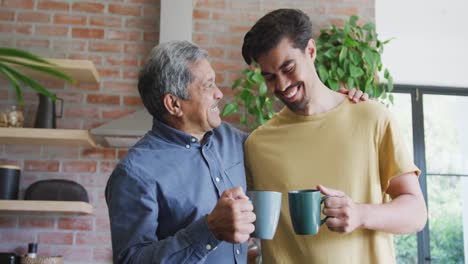 Cheerful-father-and-son-toasting-coffee-mugs-while-spending-leisure-time-in-kitchen-at-home