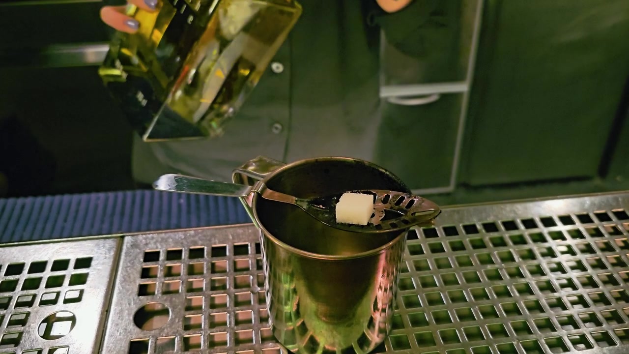 Rituale tradizionale dell'assenzio in un bar in penombra, con zolletta di zucchero su cucchiaino forato, tazza di metallo, bottiglia verde, attrezzi da barista e superficie riflettente in acciaio inossidabile sotto un'illuminazione colorata.