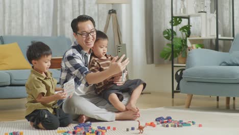 cuerpo lleno de padre y hijos asiáticos jugando el conjunto de construcción ladrillo de juguete de plástico colorido en casa. sonriendo y saludando al teléfono inteligente, haciendo videollamas, pasando tiempo juntos