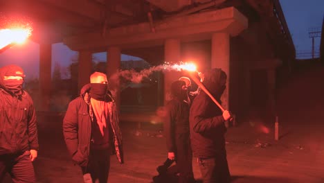 group of young men in balaclavas with red burning signal flare walking on the road under the bridge, slow motion