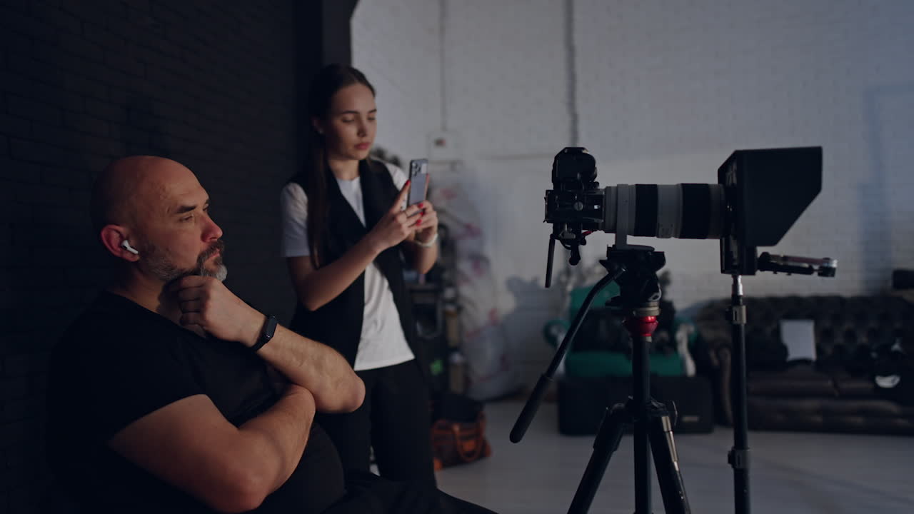 Calm relaxed Caucasian cameraman sits on the chair rubbing his beard. Photographer looks at camera on tripod. Brunette woman stands behind taking video on her phone.