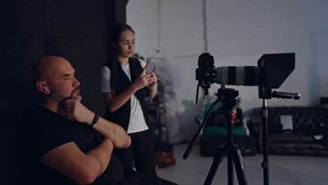 calm relaxed caucasian cameraman sits on the chair rubbing his beard. photographer looks at camera on tripod. brunette woman stands behind taking video on her phone.
