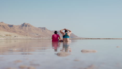 Dos-Chicas-Sentadas-En-El-Agua-Del-Mar-Muerto-Mientras-Una-Se-Arregla-El-Pelo-Con-Una-Vista-Espectacular-Y-Montañosa