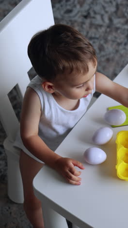lovely toddler boy sits at white desk. cute child plays with toy eggs and puzzles. top view. vertical video