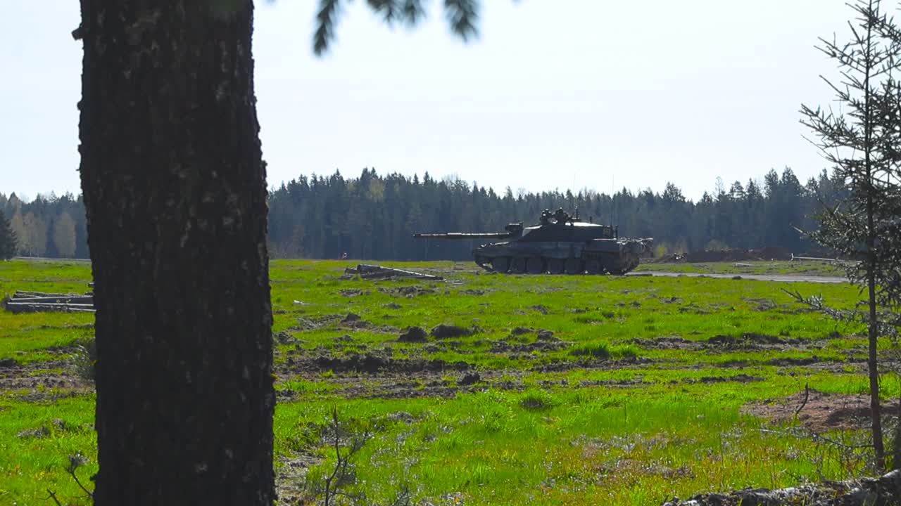 Rare vibrant footage of a British Challenger 2 4034 army tank guarding a forest road in a grassy green field during a sunny day. Filmed from a forest behind some trees and branches that are moving.