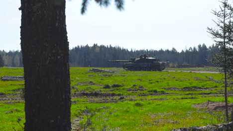 rare vibrant footage of a british challenger 2 4034 army tank guarding a forest road in a grassy green field during a sunny day. filmed from a forest behind some trees and branches that are moving.