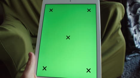 Mano-Sosteniendo-Una-Tableta-De-Pantalla-Verde-Con-Llave-De-Croma-Blanca-En-Casa-Frente-A-Un-Cómodo-Sillón-De-Fondo,-Estático