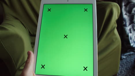 mano sosteniendo una tableta de pantalla verde con llave de croma blanca en casa frente a un cómodo sillón de fondo, estático