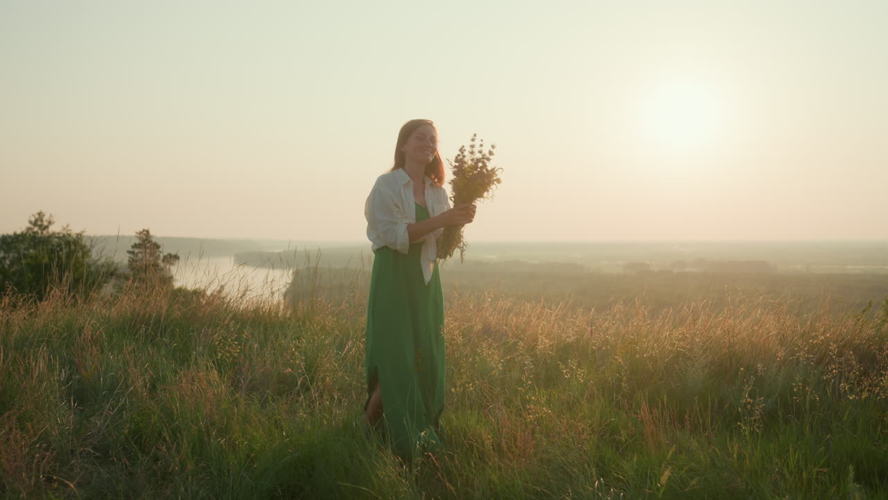 Sunlit woman in green skirt turning joyfully on grassy hilltop holding wildflower bouquet against glowing sunset sky over river valley, warm backlight highlighting silhouette