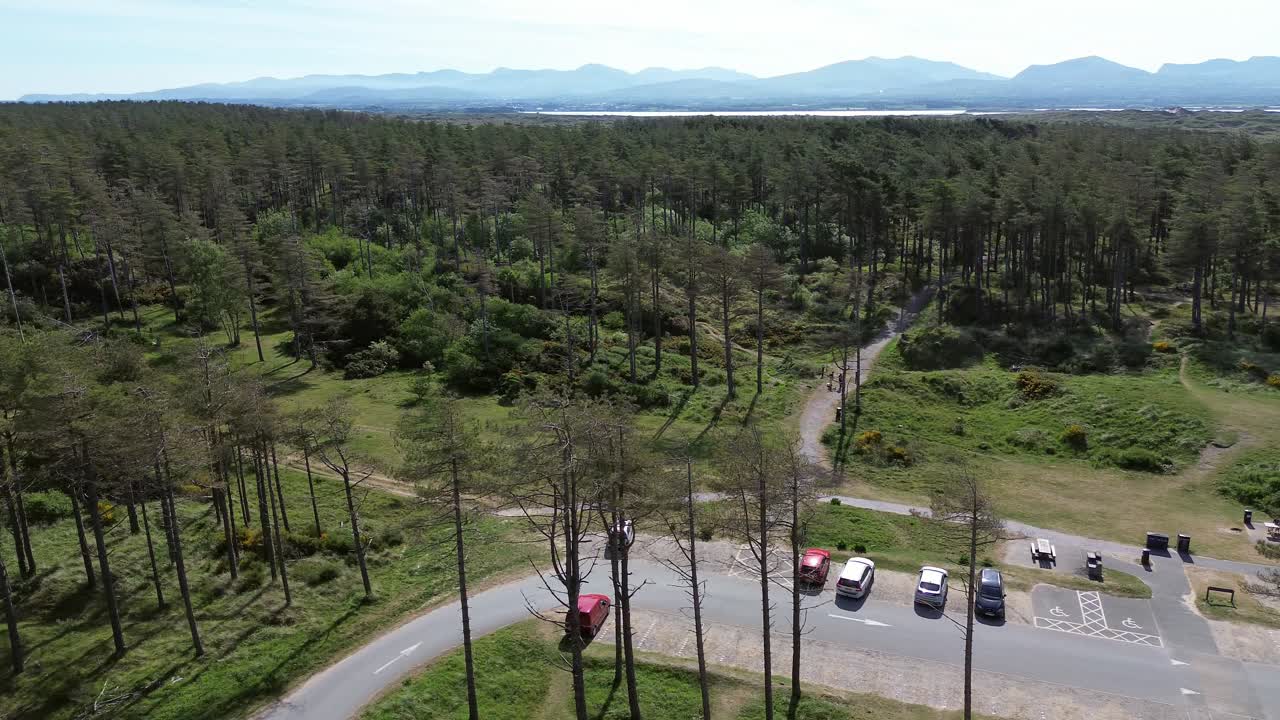 Newborough forest aerial pan view across sunny woodland car park and Snowdonia mountain range