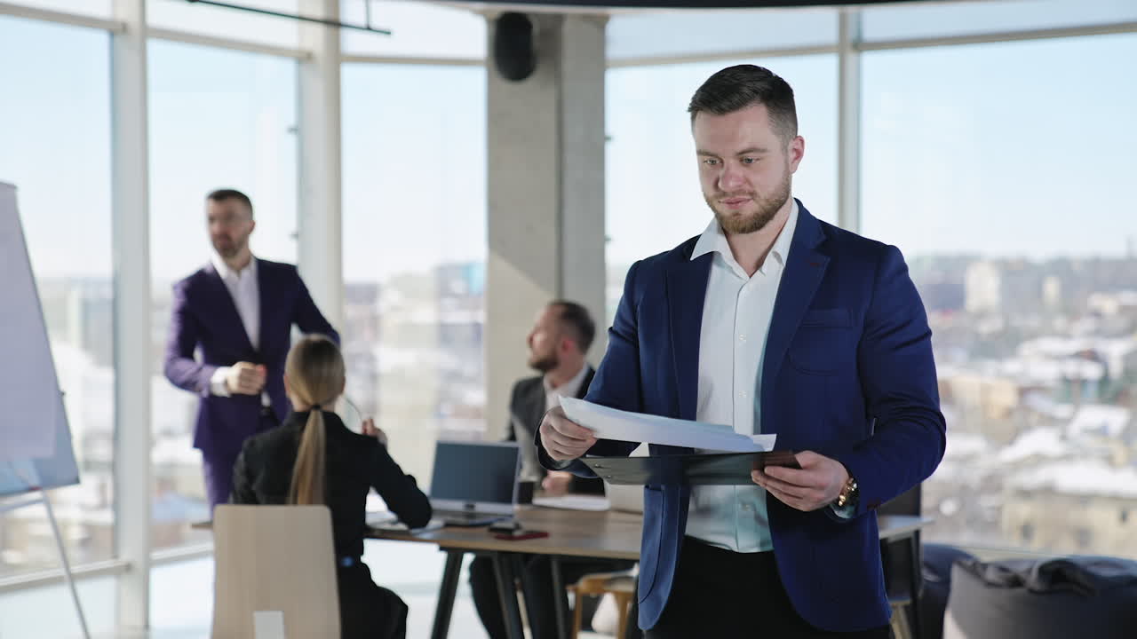 Young businessman holding documents and commenting something in front of camera. Business team working at the backdrop.