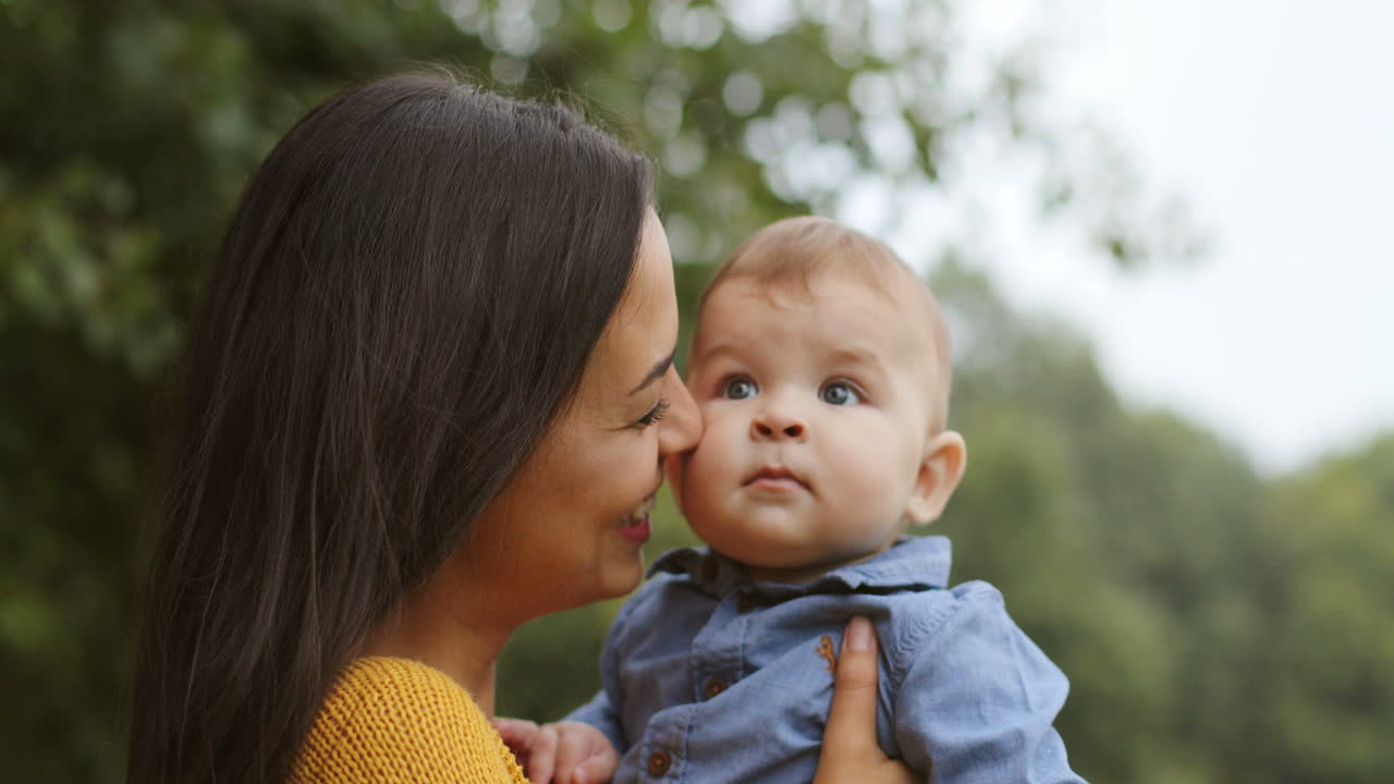cerca de una mujer feliz sosteniendo a su adorable bebé, mirándolo y besándose en el parque